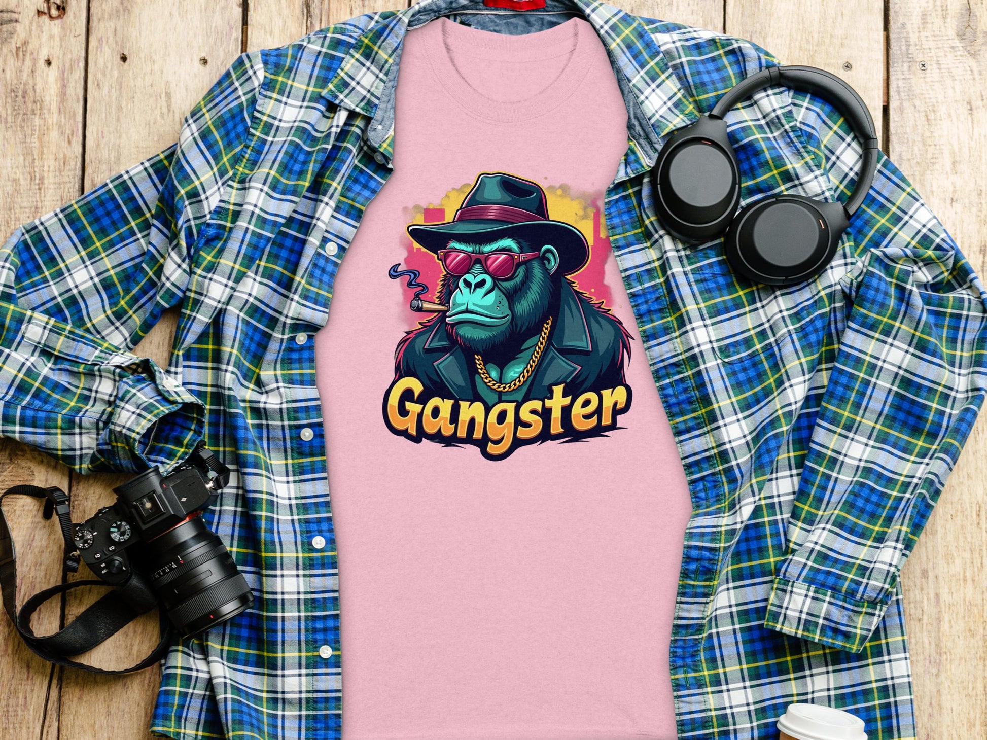 A pink Garment Graphics Gangster Gorilla T-Shirt featuring a gorilla in a hat, sunglasses, and jacket is displayed on a wooden surface alongside a classic-fit blue plaid shirt, headphones, camera, and disposable coffee cup.