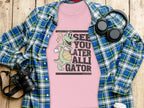 A pink t-shirt by Garment Graphics featuring a funny cartoon alligator and the phrase See you later alligator pairs perfectly with a womens blue and green plaid shirt. This outfit, set on wood, is accessorized with essentials like a camera, headphones, and a disposable cup.