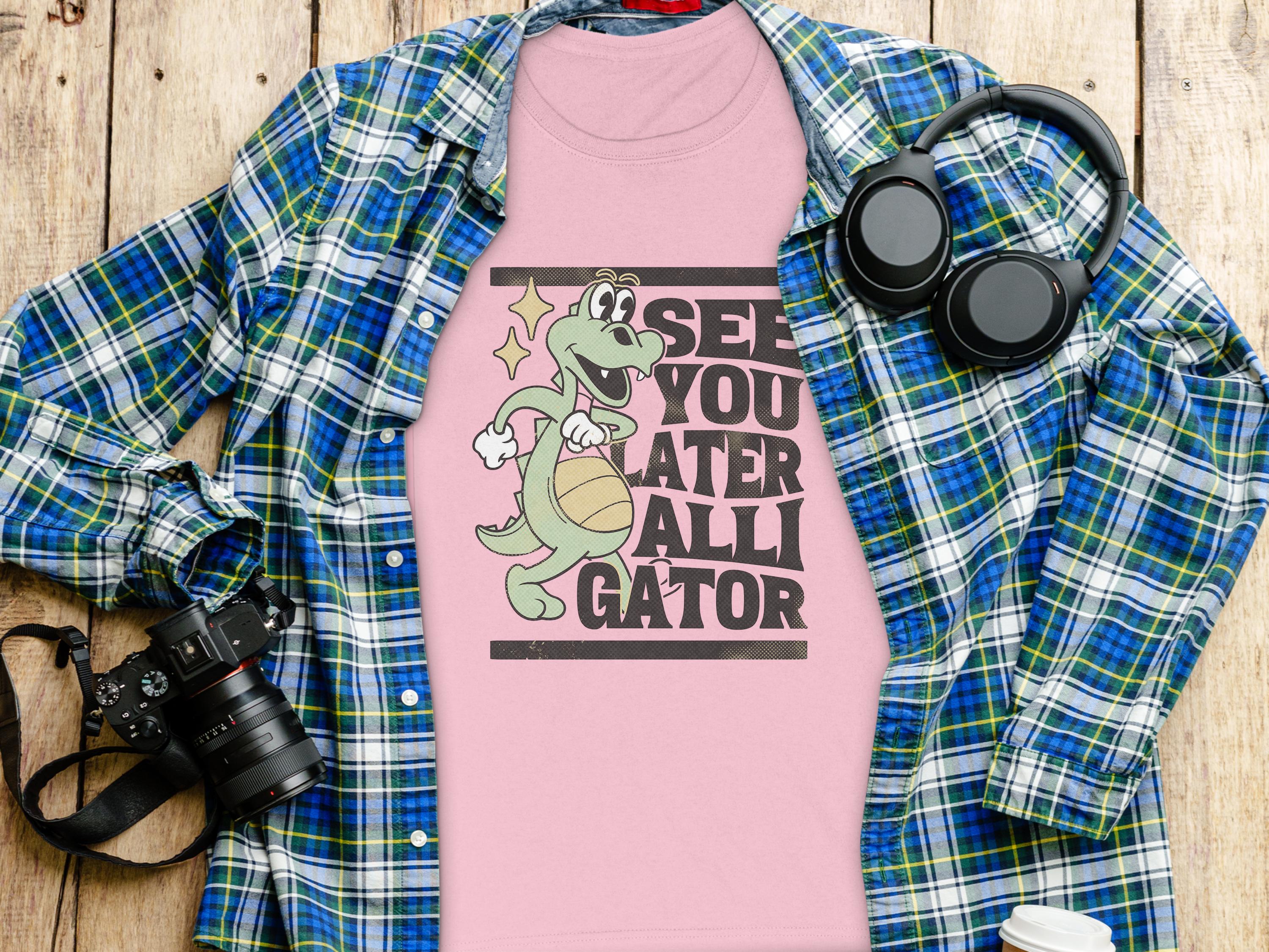 A pink t-shirt by Garment Graphics featuring a funny cartoon alligator and the phrase See you later alligator pairs perfectly with a womens blue and green plaid shirt. This outfit, set on wood, is accessorized with essentials like a camera, headphones, and a disposable cup.