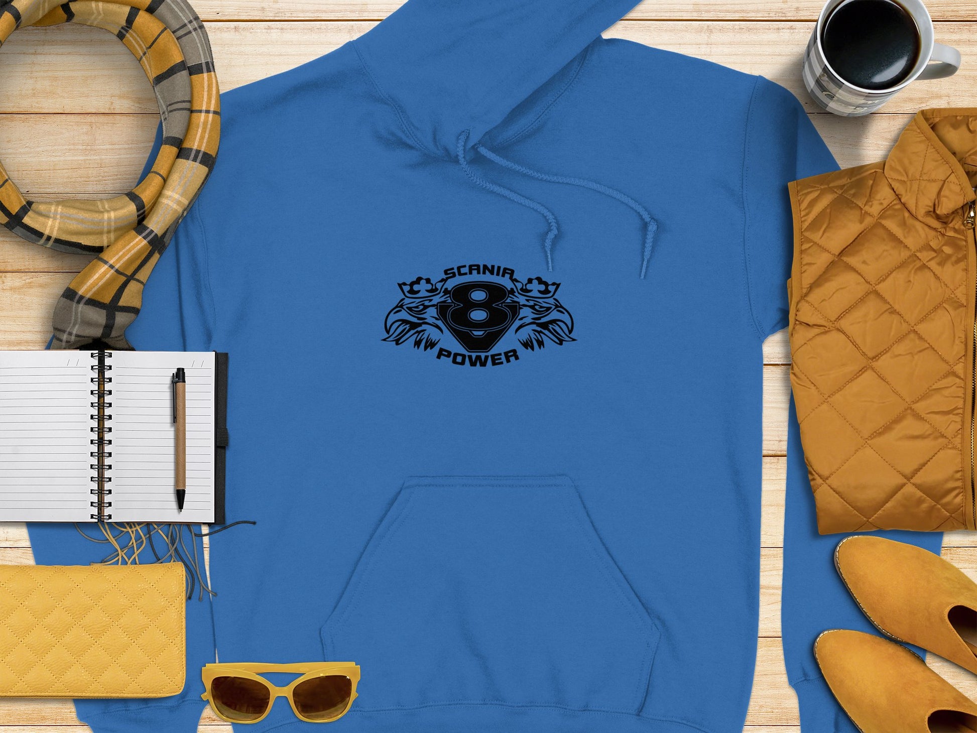 A Garment Graphics Scania Power Eight Hoodie, classic fit with Scania V8 Power and bold black engine graphics, rests on wood. Nearby items include a yellow quilted jacket, purse, notebook, pen, cup, scarf, sunglasses, and smartphone.