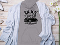 Garment Graphics gray hoodie with a classic fit has a black car graphic and the text ENJOY Classic CAR, Vintage Ride Community, Aircooled. Styled on wood with a light gray cardigan, white flowers, jewelry, and silver shoes. Ideal for fans of community spirit.