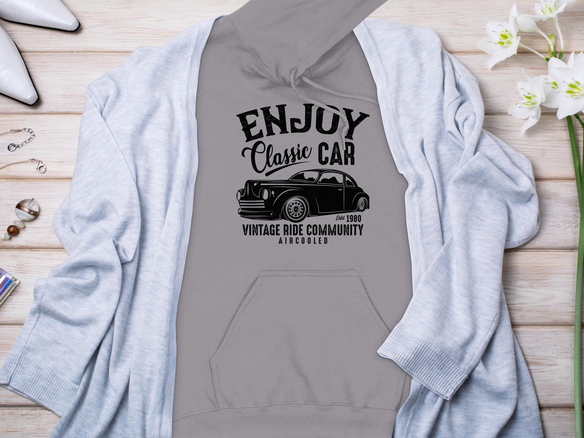 Garment Graphics gray hoodie with a classic fit has a black car graphic and the text ENJOY Classic CAR, Vintage Ride Community, Aircooled. Styled on wood with a light gray cardigan, white flowers, jewelry, and silver shoes. Ideal for fans of community spirit.