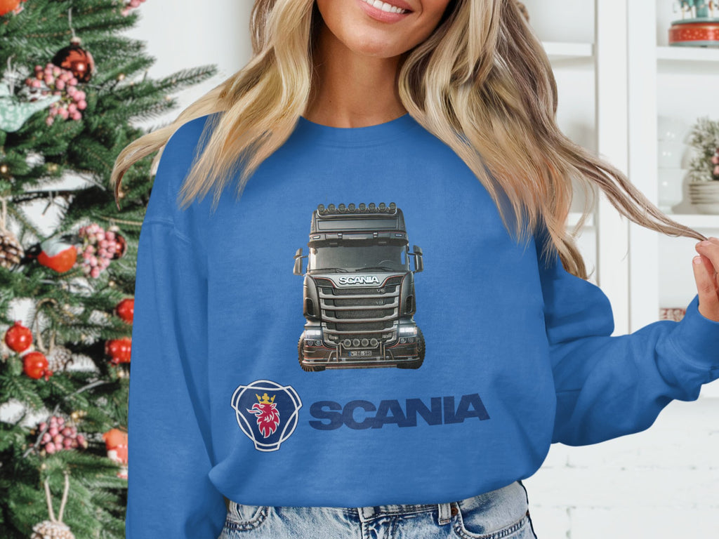 Standing indoors beside a decorated Christmas tree, theyre smiling warmly and holding a strand of hair. They wear Garment Graphics blue crewneck sweatshirt featuring the Scania truck logo—an ideal choice for truck enthusiasts.