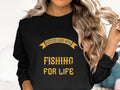 A person with long, wavy blonde hair models a Garment Graphics black Graphic Sweatshirt featuring yellow Father and Son, Fishing for Life text. They hold a strand of hair, partially obscuring their face.