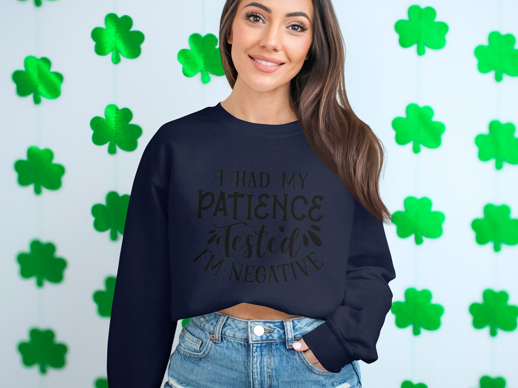 In front of a light-colored wall with green four-leaf clovers, someone wears a quirky Garment Graphics sweatshirt that says I had my patience tested, I’m negative, teamed with denim shorts—ideal for casual outings thanks to its unisex sizing.