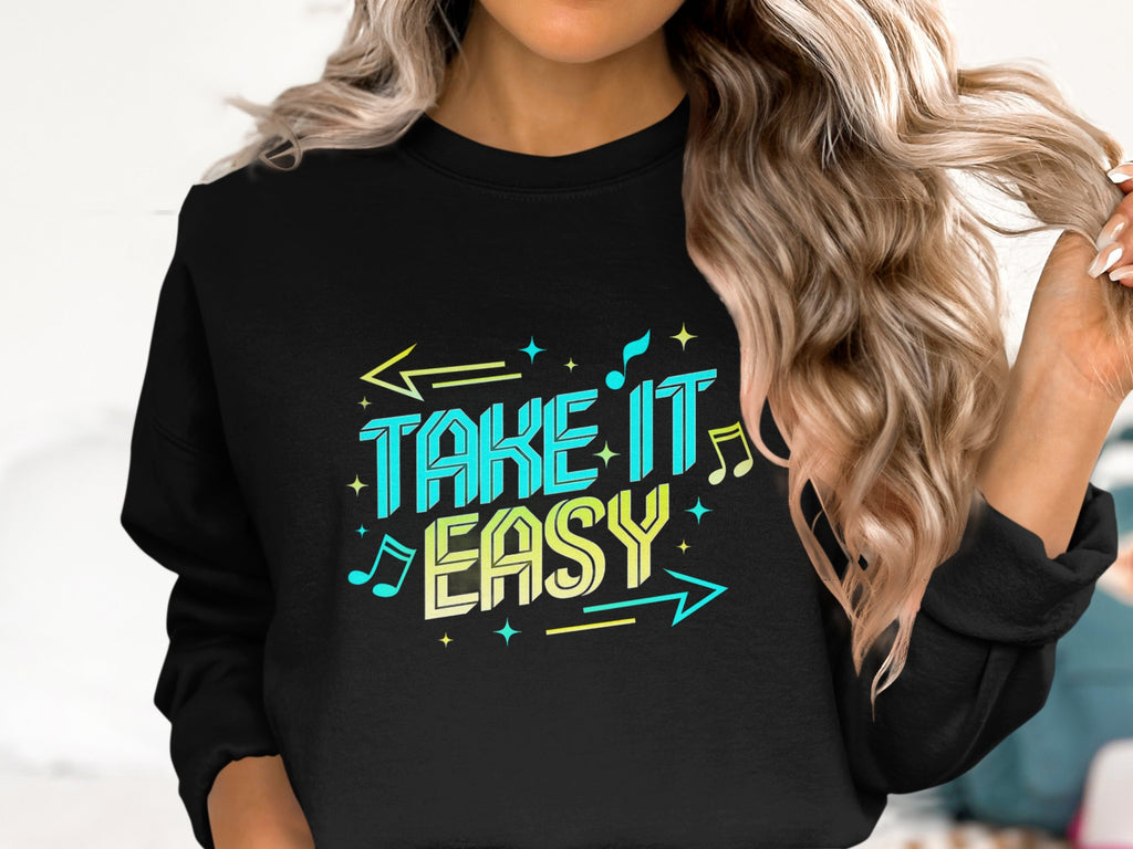 A person wears a Garment Graphics black sweatshirt with the Take It Easy design in vibrant letters, musical notes, and arrows. This comfy unisex piece perfectly accents their wavy hair, epitomizing chic casual style.