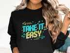A person wears a Garment Graphics black sweatshirt with the Take It Easy design in vibrant letters, musical notes, and arrows. This comfy unisex piece perfectly accents their wavy hair, epitomizing chic casual style.