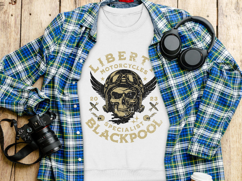 A Garment Graphics white sweatshirt with a unique Liberty Motorcycles skull design is on a wooden surface, surrounded by a blue plaid shirt, black camera, and headphones. The bold graphic reads, Liberty Motorcycles Specialist Blackpool 2023.
