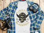 A Garment Graphics white sweatshirt with a unique Liberty Motorcycles skull design is on a wooden surface, surrounded by a blue plaid shirt, black camera, and headphones. The bold graphic reads, Liberty Motorcycles Specialist Blackpool 2023.