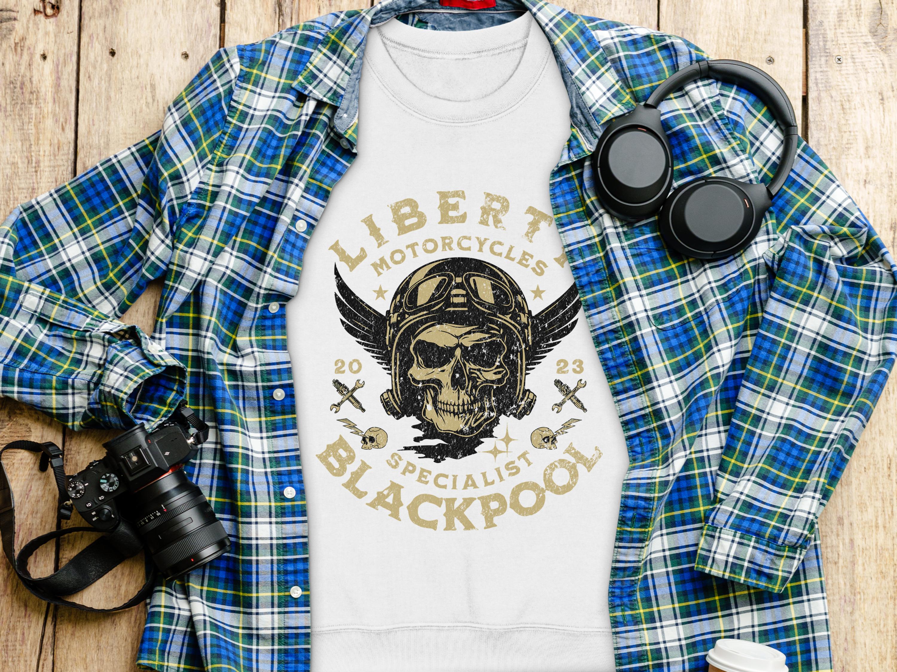 A Garment Graphics white sweatshirt with a unique Liberty Motorcycles skull design is on a wooden surface, surrounded by a blue plaid shirt, black camera, and headphones. The bold graphic reads, Liberty Motorcycles Specialist Blackpool 2023.