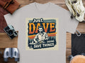 The Garment Graphics classic fit graphic T-shirt in gray features a man at a table surrounded by tools and papers, with the slogan Just DAVE doing DAVE things. Its displayed on a wooden surface with items like a camera, binoculars, gloves, and watch.