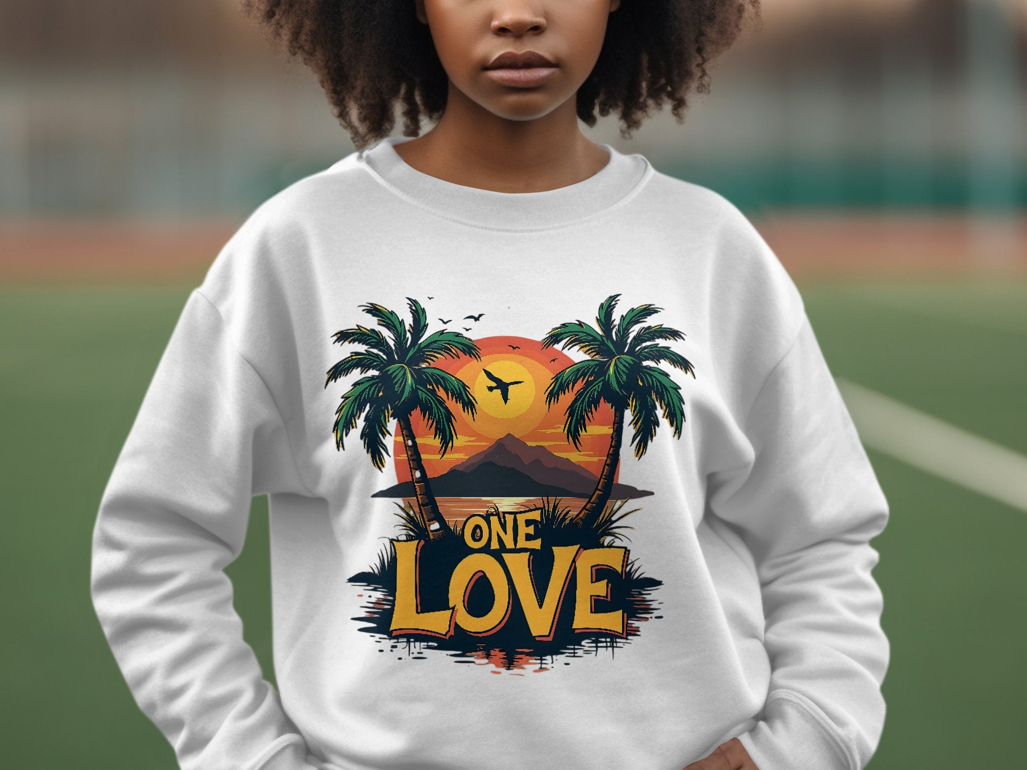 In front of a blurred backdrop, someone wears a vibrant white sweatshirt from Garment Graphics. This piece features a tropical design with palm trees and the stunning One Love Sunset, embodying the essence of relaxation and warmth.