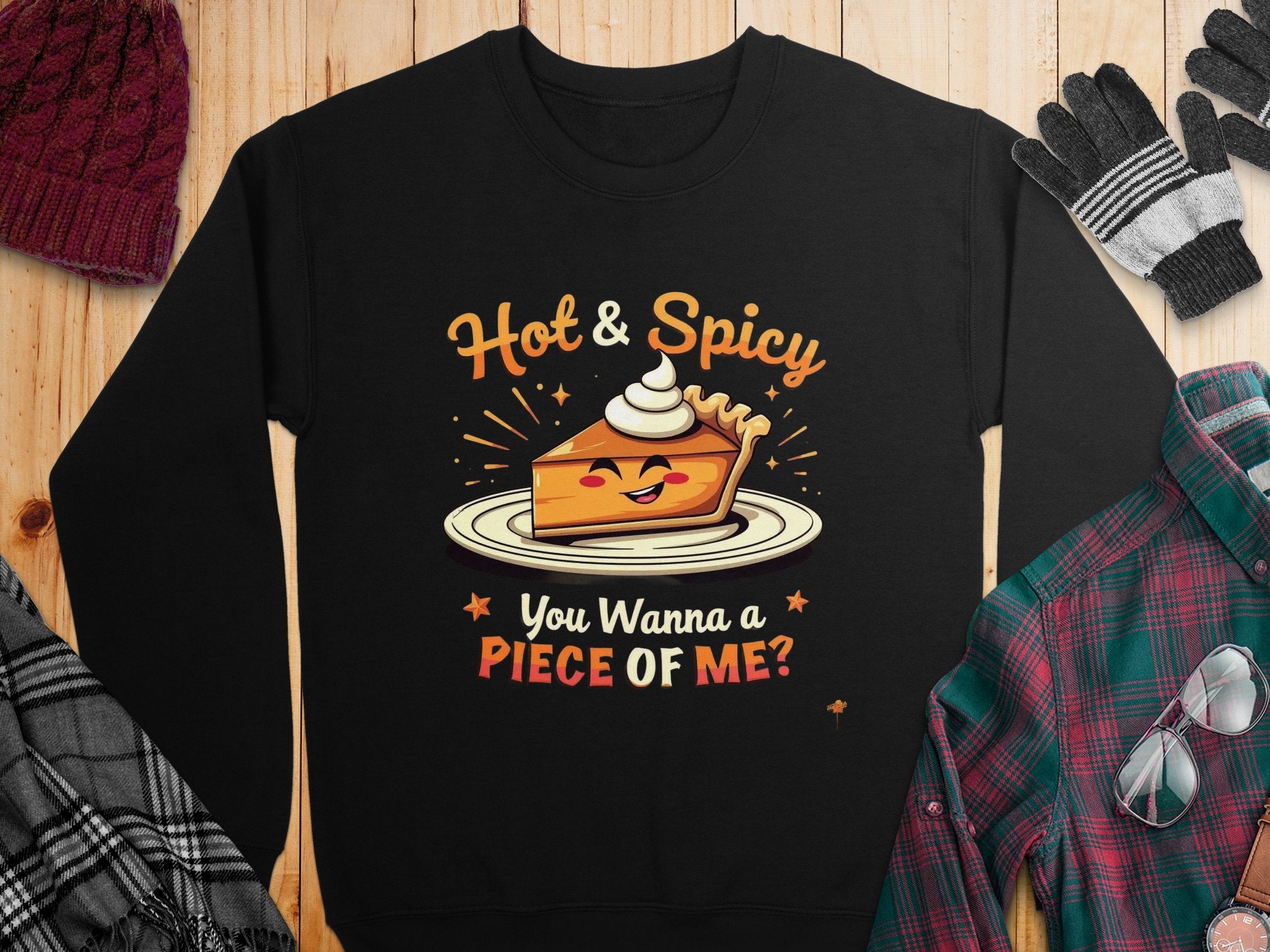 The Garment Graphics black unisex sweatshirt showcases a smiling cartoon slice of pie with whipped cream, featuring playful text Hot & Spicy and You Wanna PIECE OF ME? Perfectly paired with a hat, gloves, and plaid shirt on wood, this pie graphic sweatshirt is irresistible.