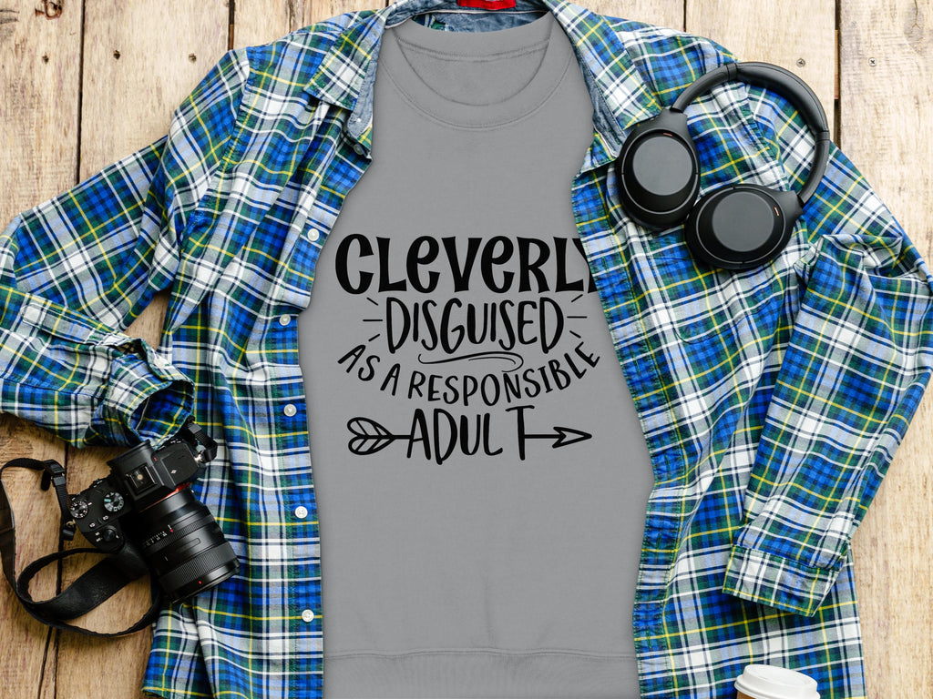 A flat lay photo showcases Garment Graphics gray t-shirt stating Cleverly Disguised As A Responsible Adult. Its surrounded by a plaid shirt, headphones, a camera, and takeout coffee on wood. Ideal for everyone with unisex sizing options!.