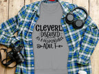 A flat lay photo showcases Garment Graphics gray t-shirt stating Cleverly Disguised As A Responsible Adult. Its surrounded by a plaid shirt, headphones, a camera, and takeout coffee on wood. Ideal for everyone with unisex sizing options!.