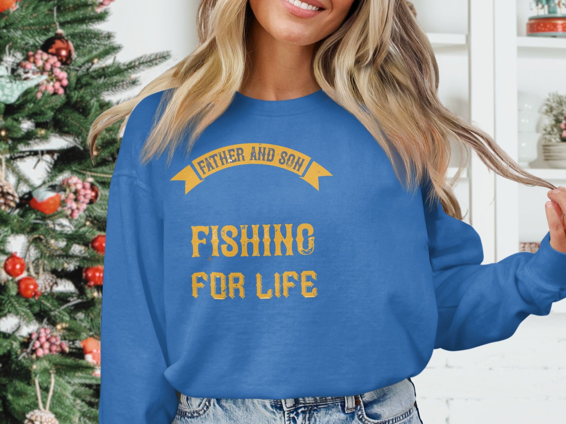 A woman wears a Garment Graphics blue graphic sweatshirt with Father and Son Fishing for Life in vibrant yellow text, standing elegantly in front of a festively decorated Christmas tree.