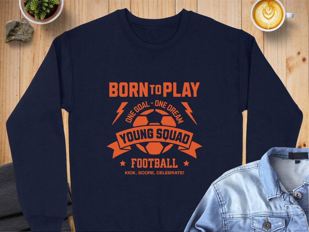 A navy blue unisex football sweatshirt from Garment Graphics features an orange graphic: Born to Play, One Goal - One Dream, Young Squad, Football, Kick, Score, Celebrate! Its displayed on a wooden table with a cappuccino, denim jacket, and small plant.