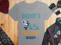 This Garment Graphics gray t-shirt features Daddys Fishing Buddy in blue and orange, depicting a fisherman and a large fish. Its an ideal womens fishing shirt, styled on wood with a red hat, gloves, and plaid shirt for added rugged charm.