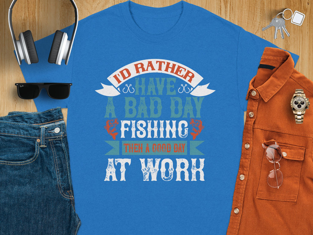 Garment Graphics Blue fishing T-shirt features Id rather have a bad day fishing than a good day at work in multicolored lettering. Ideal for anglers, it’s styled with blue jeans, orange button-up shirt, sunglasses, headphones, and watch on wood. Excellent gift choice!.