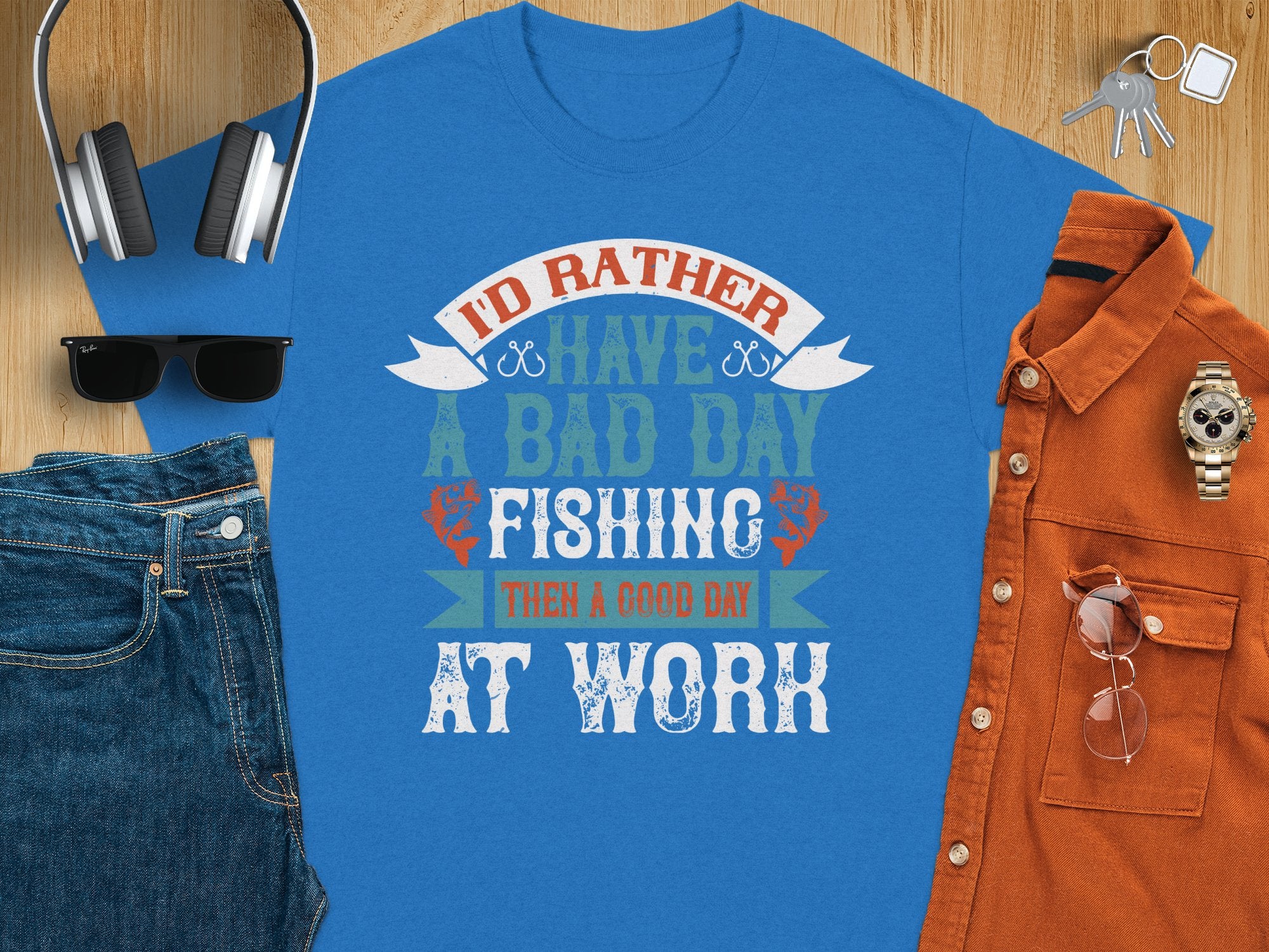 Garment Graphics Blue fishing T-shirt features Id rather have a bad day fishing than a good day at work in multicolored lettering. Ideal for anglers, it’s styled with blue jeans, orange button-up shirt, sunglasses, headphones, and watch on wood. Excellent gift choice!.
