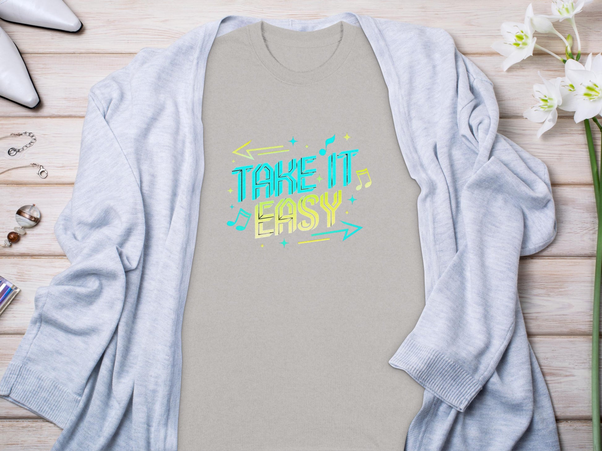 A Take It Easy shirt with colorful musical notes by Garment Graphics lies on a wooden surface, surrounded by a light gray cardigan, white flowers, jewelry, and high-heeled shoes.
