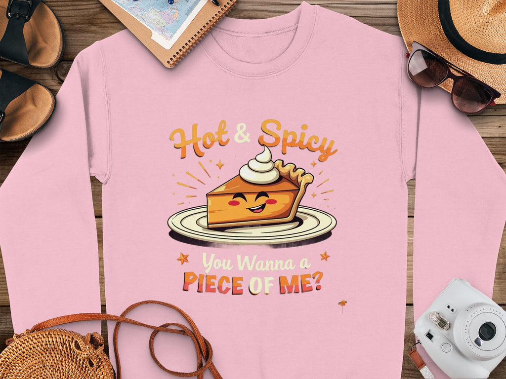 A pink Garment Graphics sweatshirt, featuring a smiling pie slice and the text Hot & Spicy You Wanna a Piece of Me? rests on wood near a camera, sunglasses, hat, shoes, and notebook. Perfect as a unisex option!.