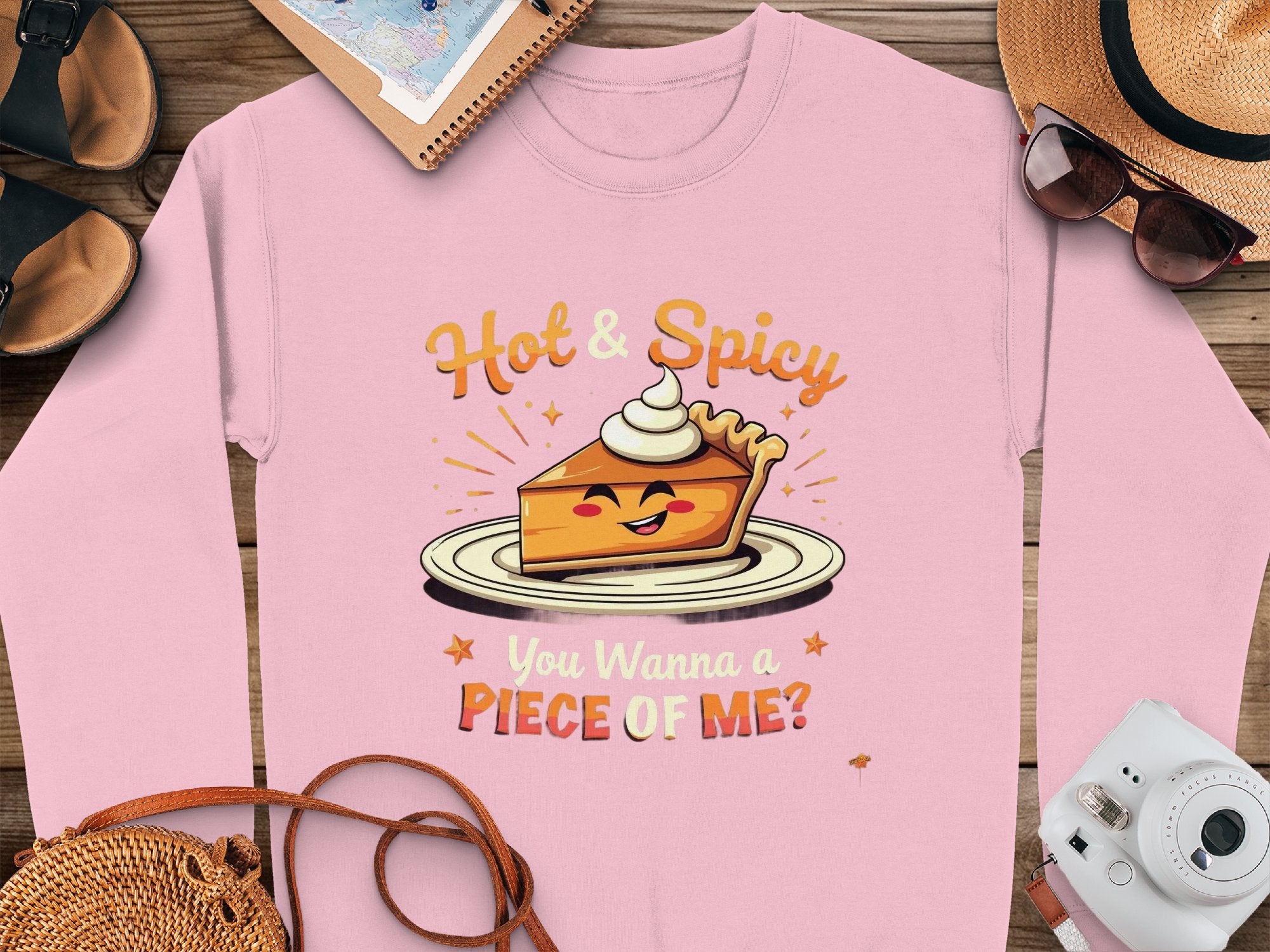 A pink Garment Graphics sweatshirt, featuring a smiling pie slice and the text Hot & Spicy You Wanna a Piece of Me? rests on wood near a camera, sunglasses, hat, shoes, and notebook. Perfect as a unisex option!.