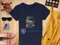 A navy blue Garment Graphics Scania T-shirt ideal for automotive fans lies on wood, featuring a bold truck design. Its surrounded by pink shoes, sunglasses, makeup brushes, a yellow blazer, nail polish, and an eyeshadow palette—an eclectic collection for the fashion-forward.