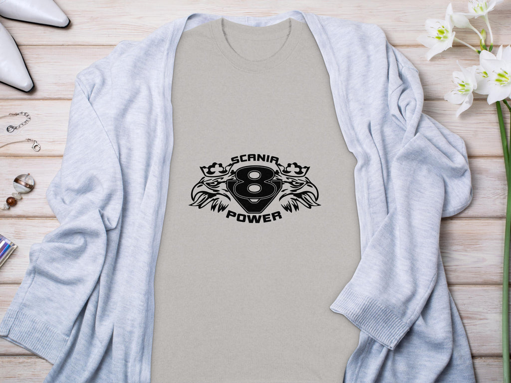 A Garment Graphics light T-shirt featuring a striking Scania Power Eagle design with two lions is laid on wood. Nearby are white heels, a gray sweater, and a vase of white flowers.