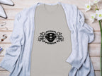 A Garment Graphics light T-shirt featuring a striking Scania Power Eagle design with two lions is laid on wood. Nearby are white heels, a gray sweater, and a vase of white flowers.