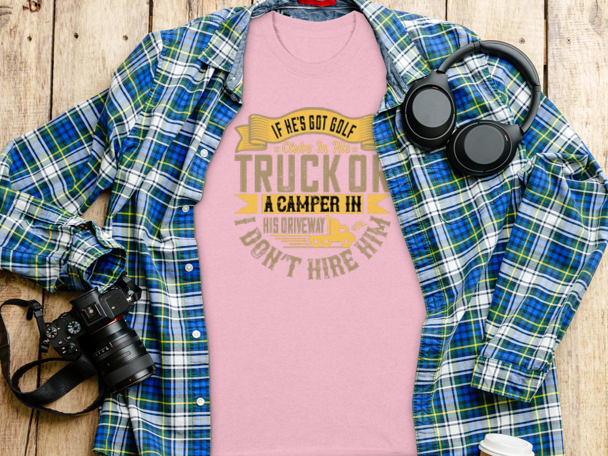A pink Garment Graphics t-shirt with If hes got golf clubs in his truck or a camper in his driveway, I dont hire him is laid on wood. Nearby are a blue plaid shirt, black headphones, and a black camera, capturing the spirit of golf and camping enthusiasts.