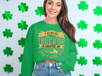 A woman in a Garment Graphics green sweatshirt featuring a humorous graphic stands against a white shamrock-decorated wall. Her long hair flows freely, smiling confidently with one hand in her pocket.