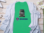 A Garment Graphics green t-shirt, showcasing a Scania truck and logo, is displayed on a wooden surface, ideal for truck enthusiasts. Its accompanied by a gray cardigan, white high heels, jewelry, and surrounded by white flowers.