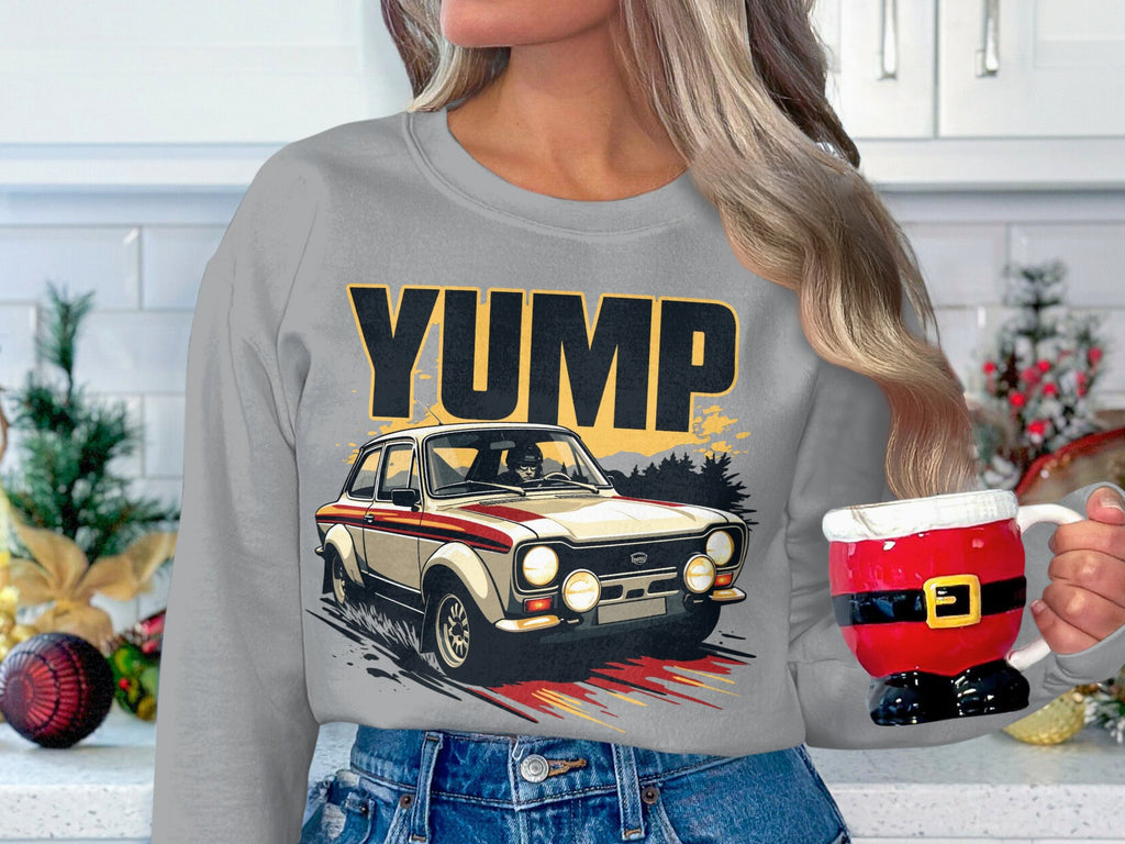 In a festive kitchen adorned with holiday decor and a white tiled backsplash, a person with long hair holds a Santa-themed mug. They are wearing a gray sweatshirt from Garment Graphics featuring the YUMP design and a rally car graphic.