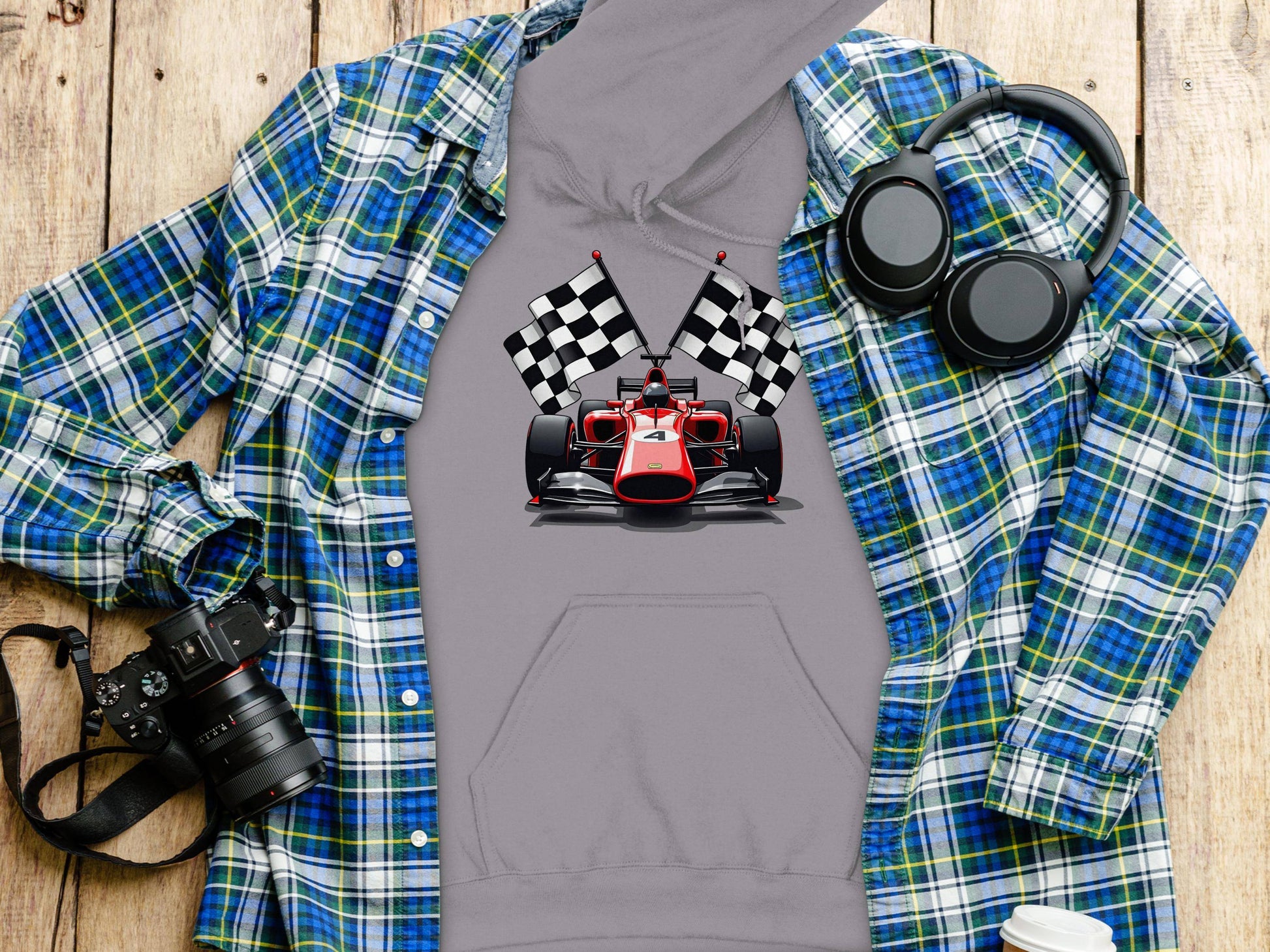 A Garment Graphics grey hoodie with a racing car and checkered flags design is stylishly displayed with a blue and green plaid shirt, over-ear headphones, a camera, and a white takeaway cup on the wooden surface.