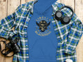 Displayed on a wooden surface are a blue plaid shirt, distinct Blackpool Hoodies from Garment Graphics featuring a skull design and Liberty Motorcycles Specialties Blackpool text, headphones, a camera, and a lidded cup.