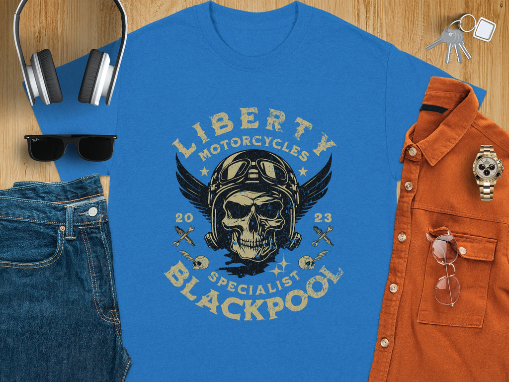 The Garment Graphics blue t-shirt features a skull with goggles and a helmet, wings on each side, and reads Liberty Motorcycles Specialist Blackpool 2023. Ideal for motorcycle enthusiasts, it pairs well with jeans, headphones, sunglasses, a watch, a brown shirt, and keys.