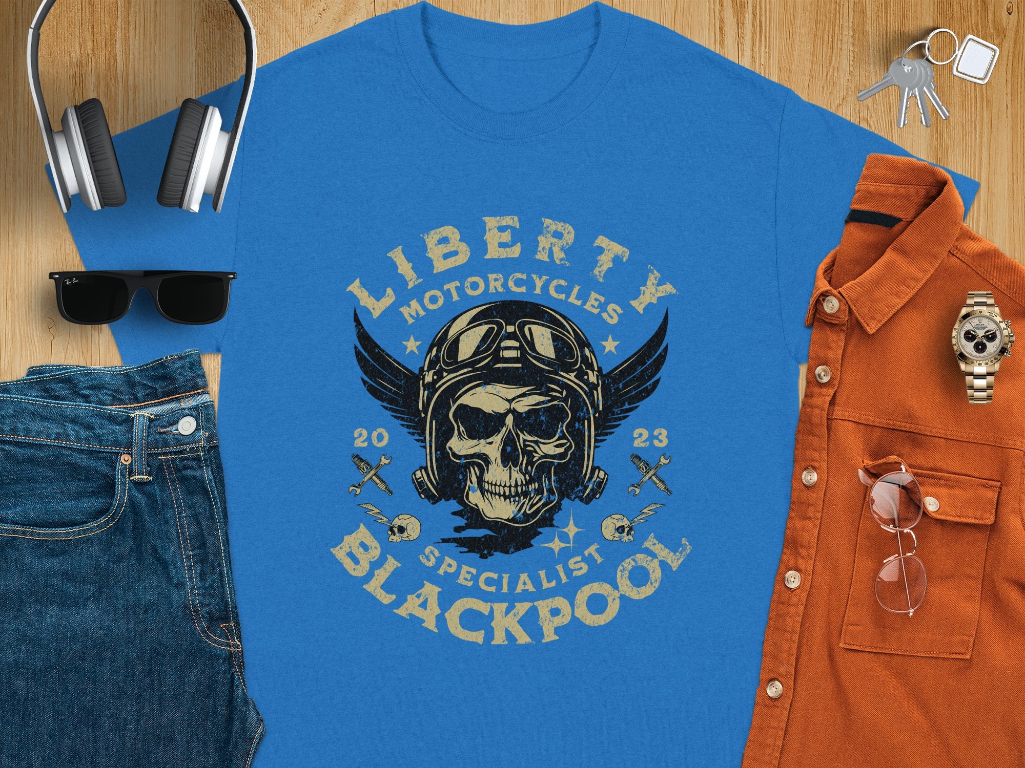 The Garment Graphics blue t-shirt features a skull with goggles and a helmet, wings on each side, and reads Liberty Motorcycles Specialist Blackpool 2023. Ideal for motorcycle enthusiasts, it pairs well with jeans, headphones, sunglasses, a watch, a brown shirt, and keys.