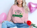 A person sits cross-legged in a festive Garment Graphics sweatshirt featuring Happy St. Patricks Day, shamrocks, and a truck graphic. They hold a red rose while pink heart-shaped balloons float behind them, enhancing the cheerful scene.