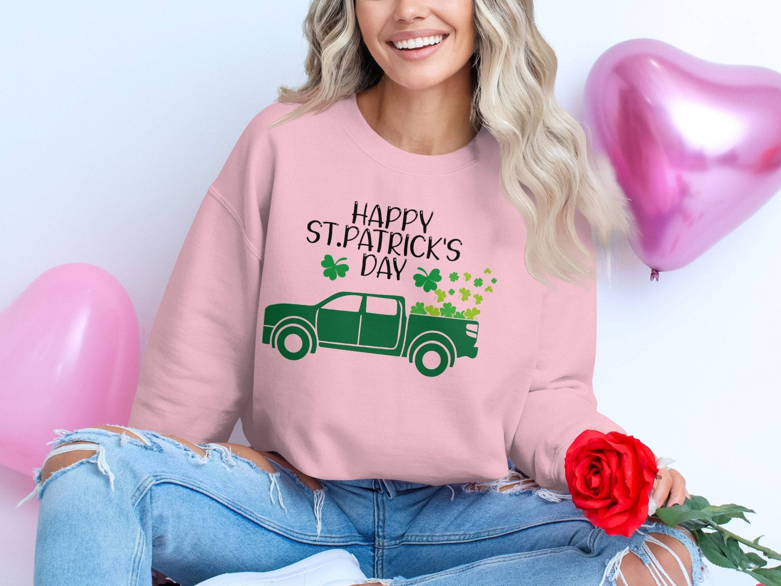 A person sits cross-legged in a festive Garment Graphics sweatshirt featuring Happy St. Patricks Day, shamrocks, and a truck graphic. They hold a red rose while pink heart-shaped balloons float behind them, enhancing the cheerful scene.
