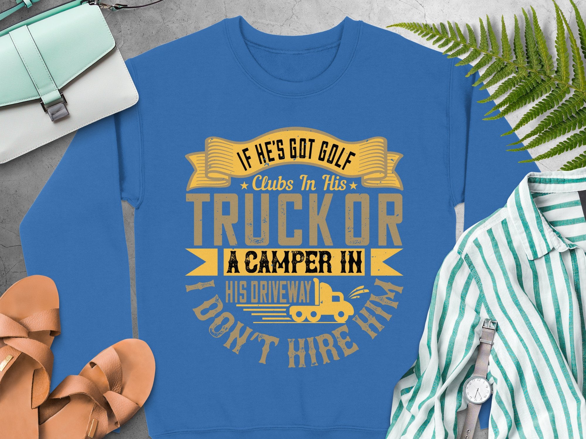Garment Graphics blue humor sweatshirt says, If hes got golf clubs in his truck or a camper in his driveway I dont hire him, with a camper graphic. Nearby are casual items like sandals, a striped shirt, ferns, and a light blue crossbody bag.