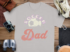 A Garment Graphics gray T-shirt with Soccer Dad in pink bubble letters above a soccer ball rests on wood, accompanied by a camera, binoculars, a leather pouch, and a wallet—ideal for the sporty dad who loves capturing every game moment.