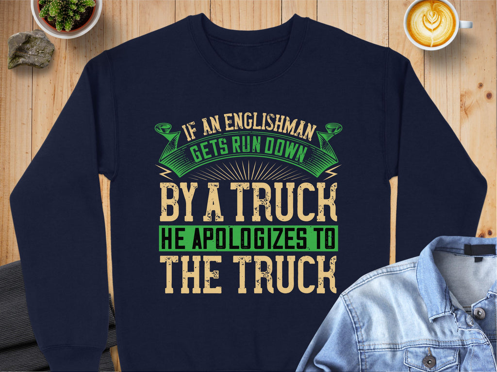 A navy blue Garment Graphics sweatshirt boasts a humorous design: If an Englishman gets run down by a truck, he apologizes to the truck, in green and beige letters. Displayed on a wooden table with a denim jacket and latte, this unisex piece is universally appealing.