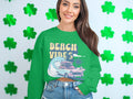 A woman with long hair stands before a shamrock-decorated white wall, wearing a green Beach Vibes sweatshirt by Garment Graphics, featuring a wave and classic car design.