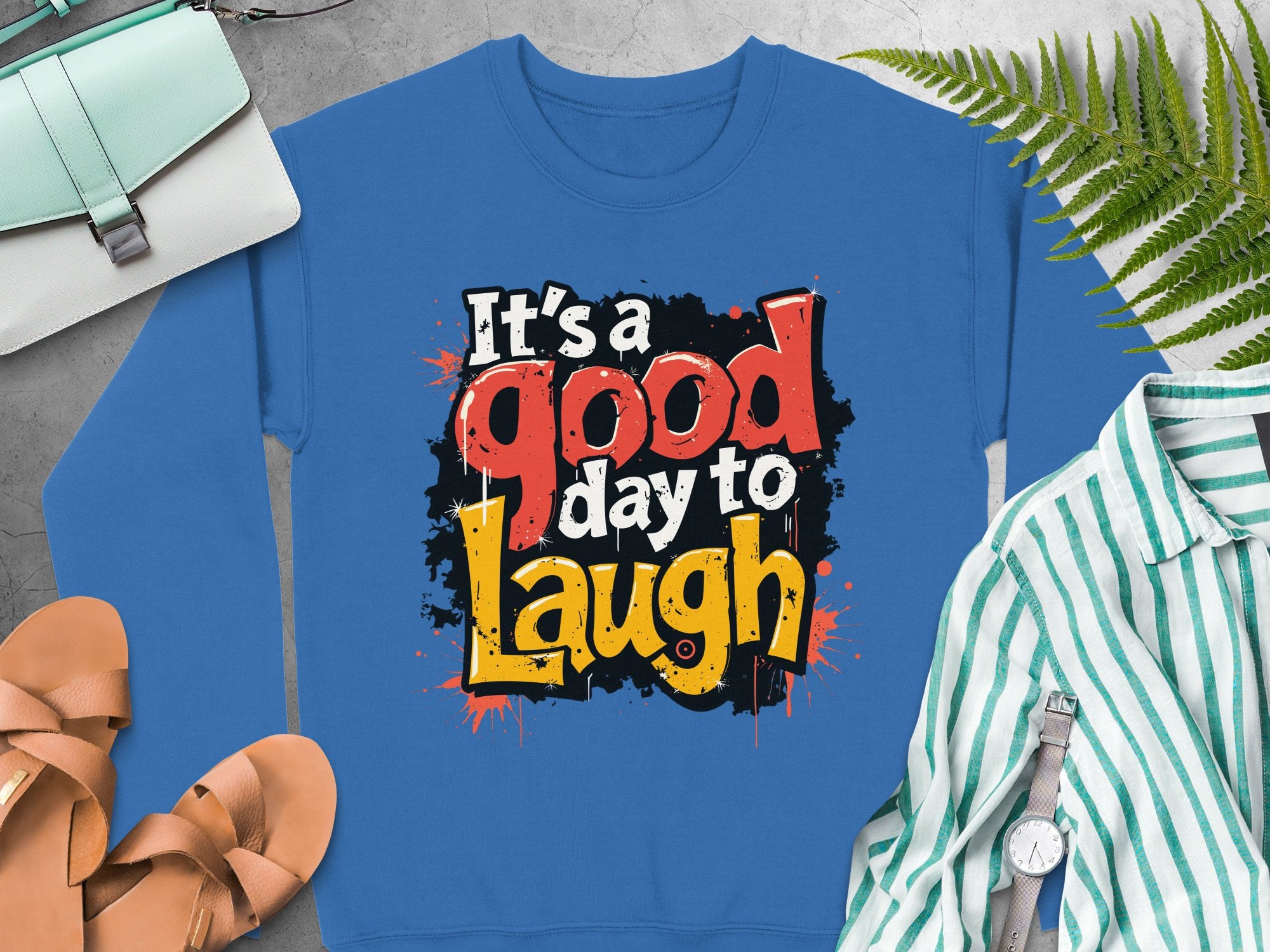 Garment Graphics offers a blue graphic printed sweatshirt featuring Its a good day to Laugh in colorful, playful fonts. The display includes a striped shirt, sandals, wristwatch, and light blue bag on a gray surface. It’s unisex-sized and made from medium-heavy fabric for comfort.