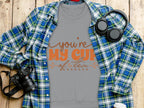 A cozy Garment Graphics sweatshirt sits beneath a gray tee with Youre My Cup of Tea in bold orange. Ideal for tea lovers, its stylishly placed on a plaid blue and green shirt, surrounded by a camera, headphones, a bottle, and coffee cup on a wooden surface.