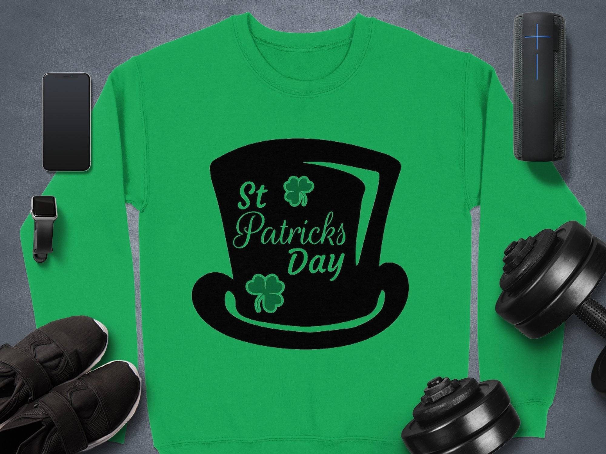 Garment Graphics presents a stylish green sweatshirt featuring a black top hat with shamrocks and St Patricks Day text, perfectly capturing the festive spirit. Its surrounded by a smartphone, smartwatch, wireless speaker, shoes, and dumbbells on a gray background.