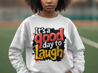 A person wears a Garment Graphics white sweatshirt with the bold, colorful phrase Its a good day to laugh. This medium-heavy fabric sweatshirt features unisex sizing and is displayed against a blurred outdoor background.