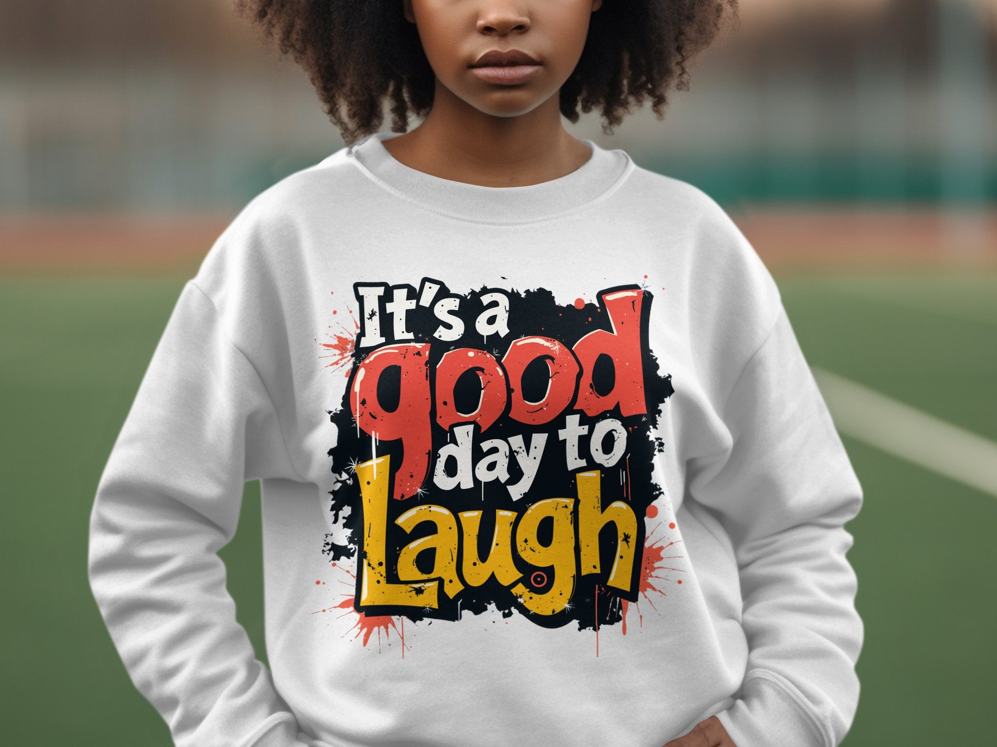 A person wears a Garment Graphics white sweatshirt with the bold, colorful phrase Its a good day to laugh. This medium-heavy fabric sweatshirt features unisex sizing and is displayed against a blurred outdoor background.