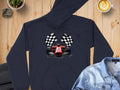 A black Garment Graphics hoodie with a red racing car and crossed checkered flags is showcased on wood alongside a denim jacket, a small plant, and a latte art coffee, all encircled by natural rocks.